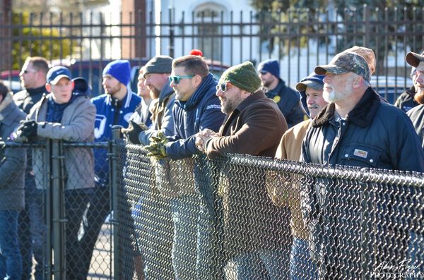 A group of men watching an event behind a chain-link fence on a sunny day.