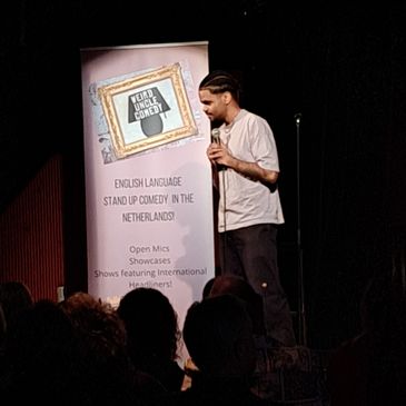 A comedian performs stand-up comedy on stage with a banner in the background.