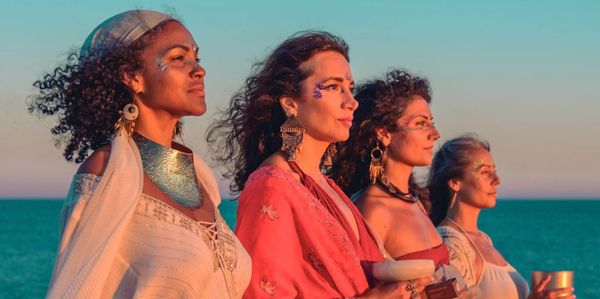 Four women in bohemian attire stand by the ocean during sunset.