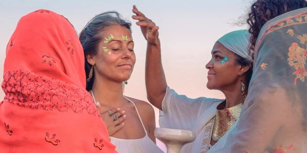 Women in colorful attire performing a spiritual ritual with face paint at sunset.