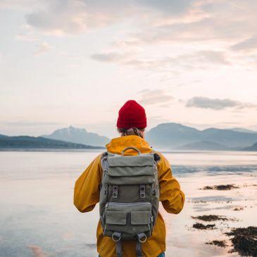 Person in red hat and yellow jacket looks at calm water and mountains. Client Journey.