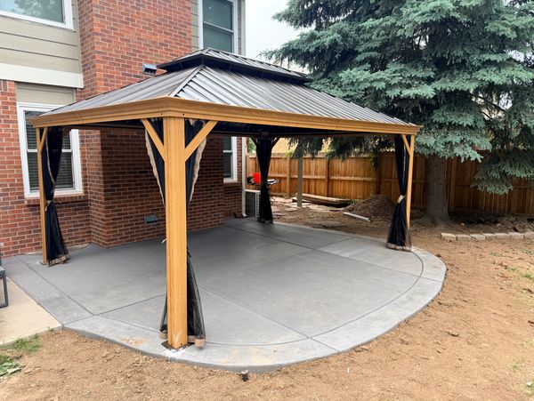 A wooden gazebo with a metal roof on a concrete patio beside a brick house.