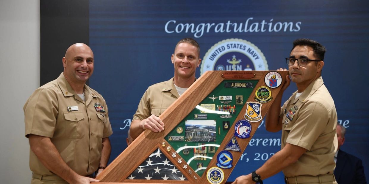 Three Navy personnel holding a decorated display case with medals and patches.