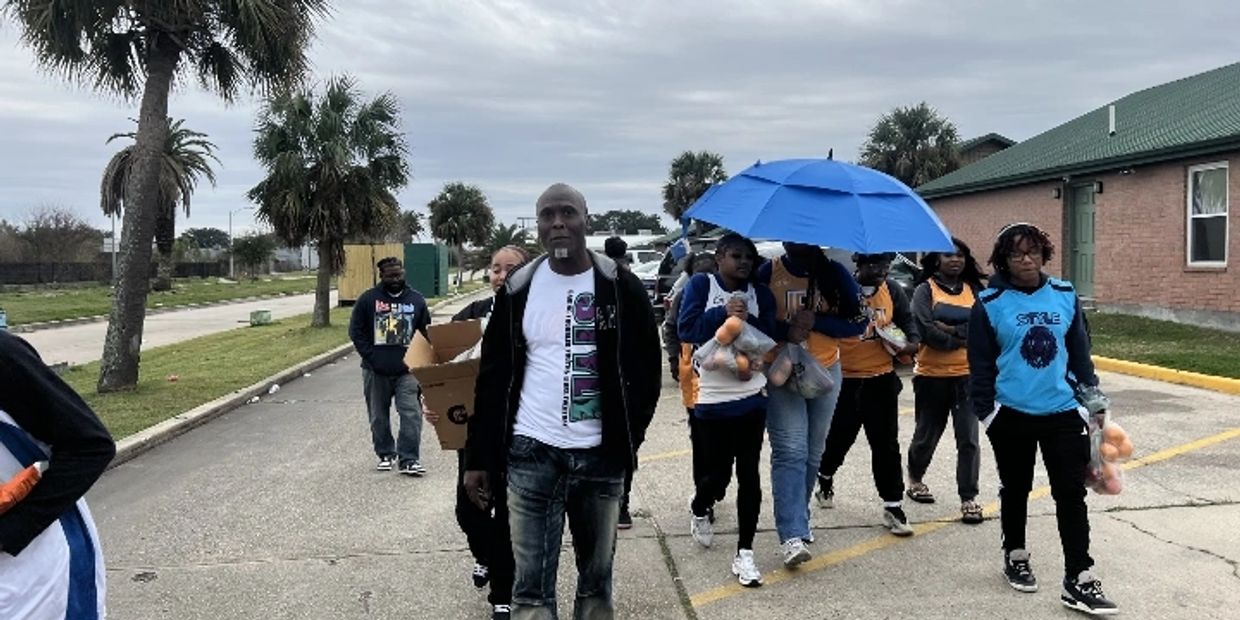 A group of people walking outdoors, some carrying bags and a blue umbrella.