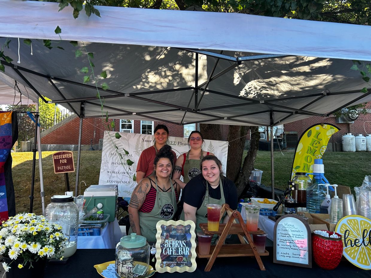 Four smiling women at a lemonade stand under a canopy.