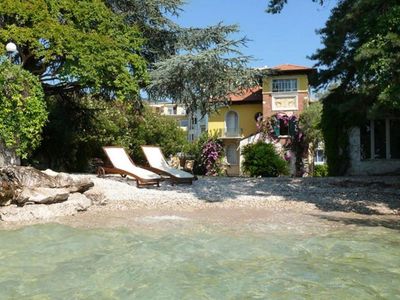 Two lounge chairs on a pebble beach with a villa and lush greenery in the background.