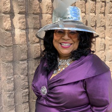 A joyful woman in a purple dress and silver hat smiles warmly.