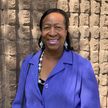 Smiling woman in a purple blazer stands against a textured brick wall.