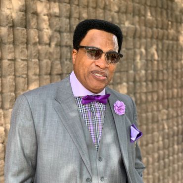 Man in a gray suit with purple accessories and sunglasses.