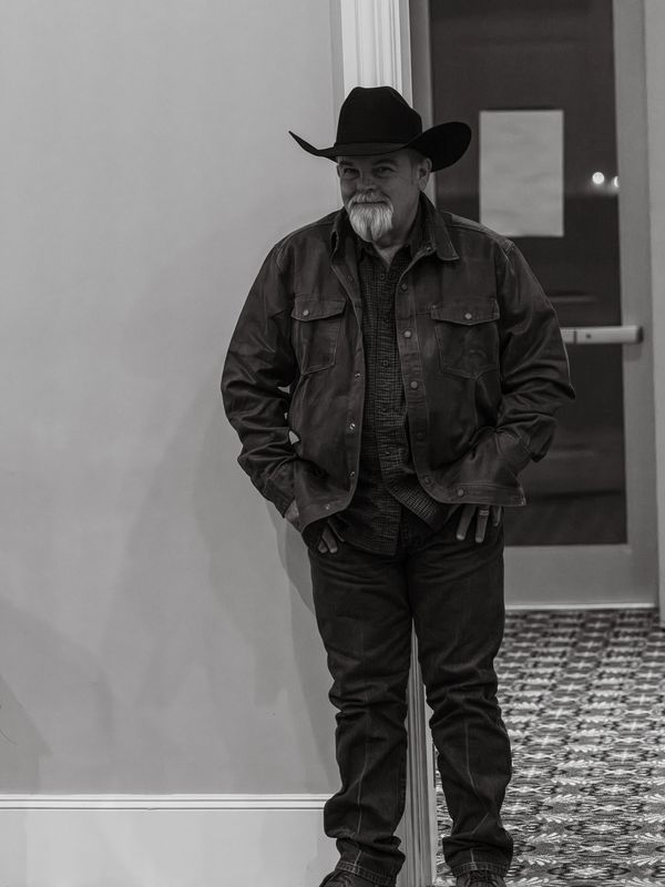 Man in cowboy hat and leather jacket leans against a wall indoors.