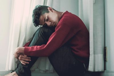 Man in red sweater sitting curled up by a window.