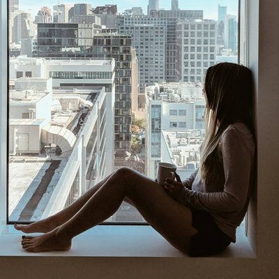 Woman sitting by window, holding a cup, overlooking city buildings.