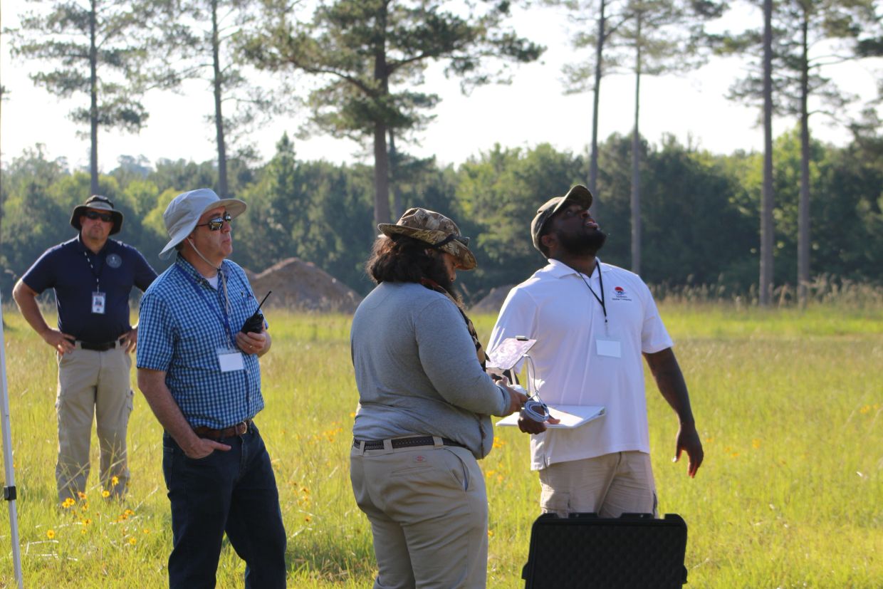 Four men in an outdoor field using a drone controller.