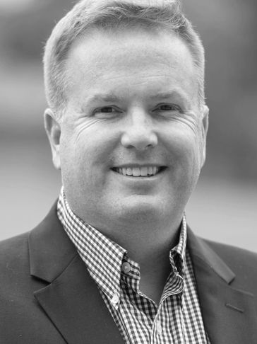 Smiling man in a checkered shirt and dark blazer outdoors.