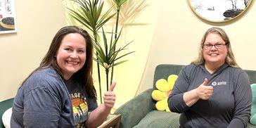Two women sitting on couches smiling and giving thumbs up signs indoors.