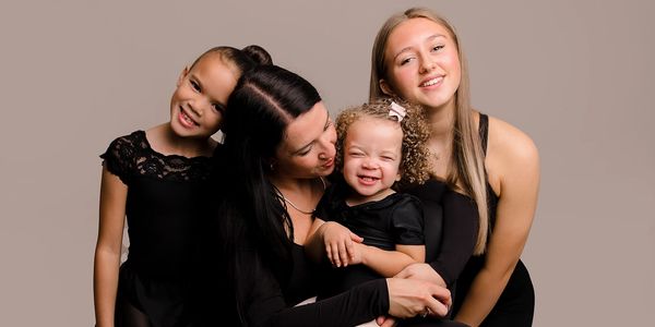 A joyful mother embraces her three smiling daughters against a neutral background.