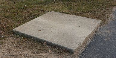 Square concrete slab embedded in the ground near grass and pavement.