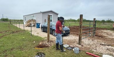 Man working on a rural fence near a mobile home and parked car.