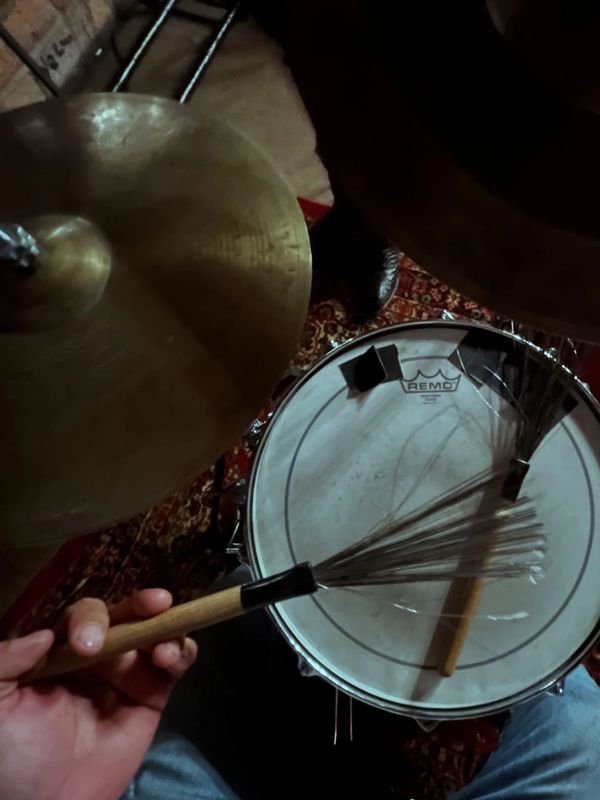 Hand holding a wire drum brush above a snare drum and cymbal.