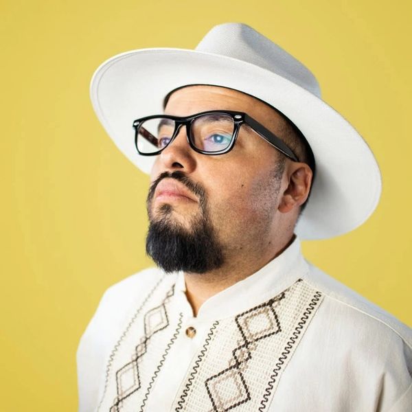 Man in white traditional shirt and hat with glasses against yellow background.