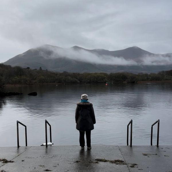 Person stands alone by a misty lake with mountains in the background.