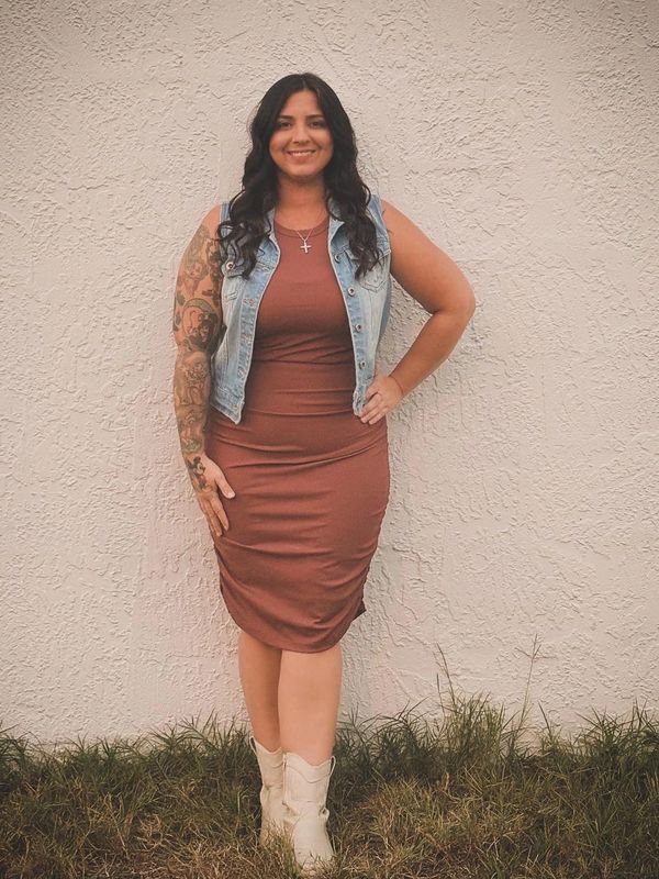 Woman in a brown dress and denim vest smiling against a textured wall.