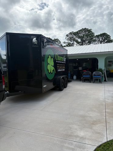 Black trailer with a green clover logo parked outside a home garage.