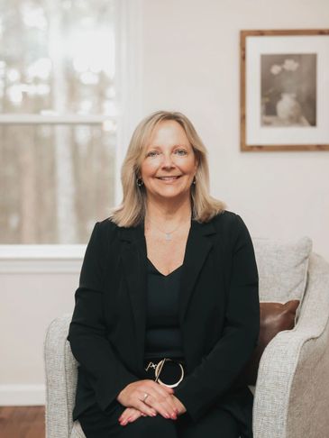 Smiling woman in black blazer sitting on a gray armchair indoors.