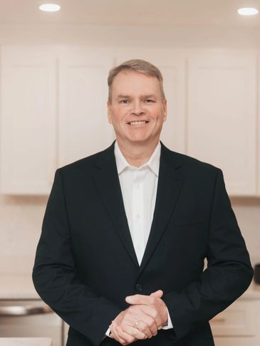 Smiling man in a black suit standing indoors with hands clasped.