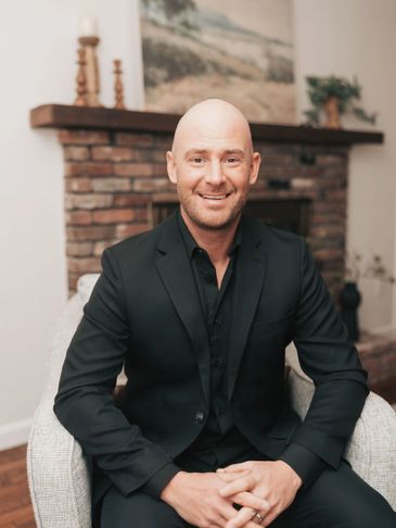 Smiling bald man in a black suit sitting in a cozy chair indoors.