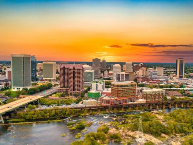 City skyline at sunset with river and greenery in the foreground.