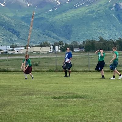 People playing with a caber in a field with mountains in the background.