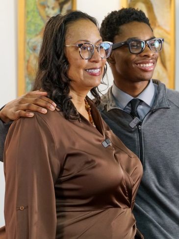A woman and young man smiling together, both wearing glasses and microphones.