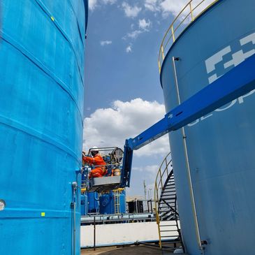 Worker in orange suit performing maintenance on large blue industrial tanks.
