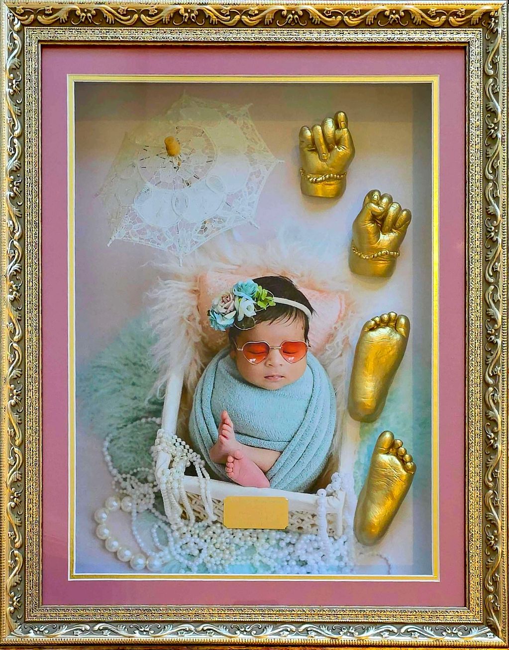 Framed baby photo with golden hand and foot molds and decorative pearls.