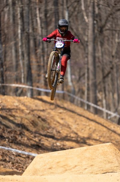 A cyclist in red jumps over a dirt ramp in the forest.