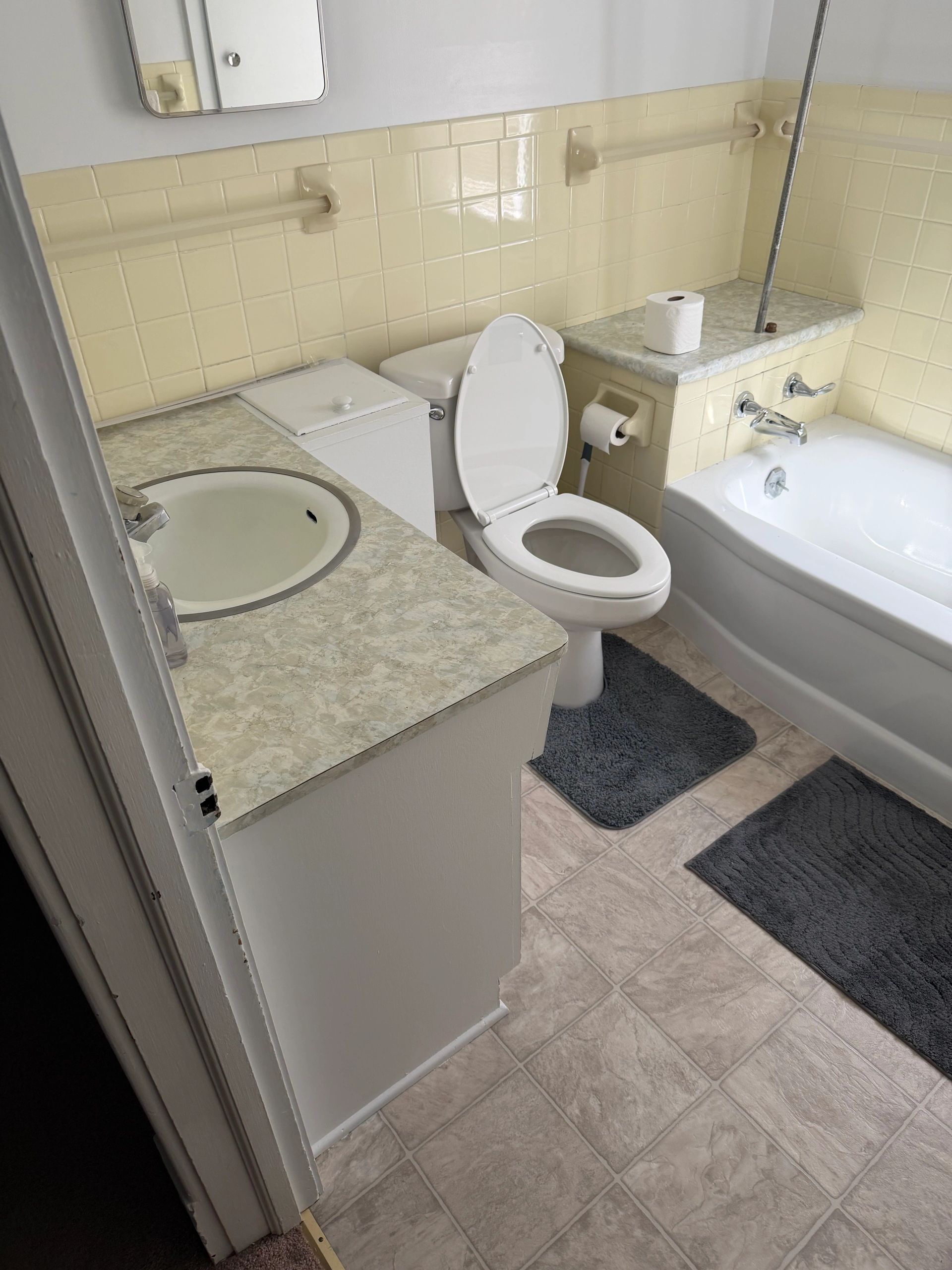 Small bathroom with yellow tiles, white fixtures, and gray rugs.