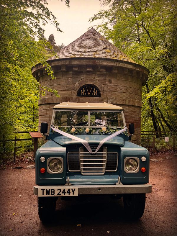 Vintage blue Land Rover decorated with white ribbons and flowers in front of a round stone building.
