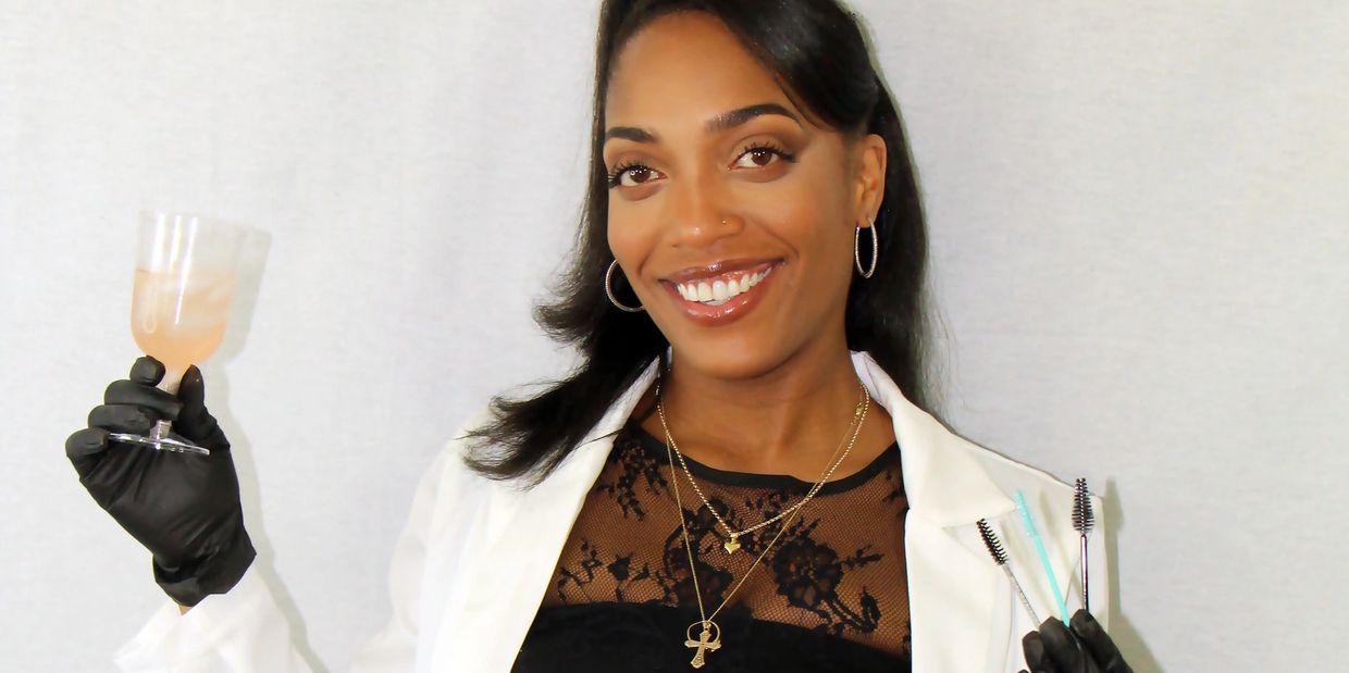 Smiling woman in white suit and black gloves holding a drink and cosmetic brushes.