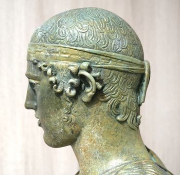 Side view of an ancient bronze statue with detailed hair and headband.