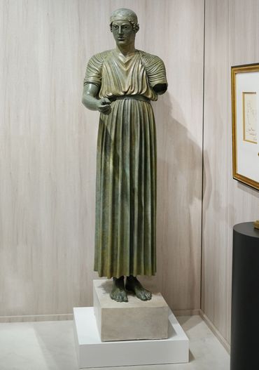 Ancient bronze statue of a robed figure missing one arm, displayed indoors on a pedestal.