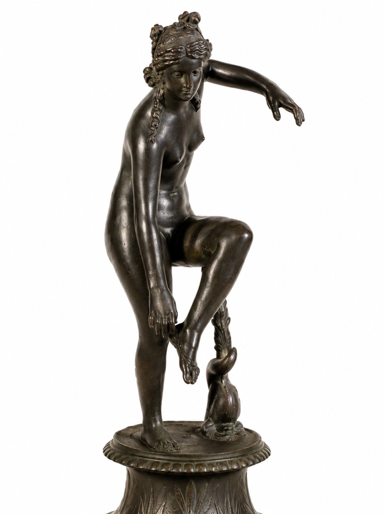 Bronze statue of a woman adjusting her sandal, standing on a decorated base.