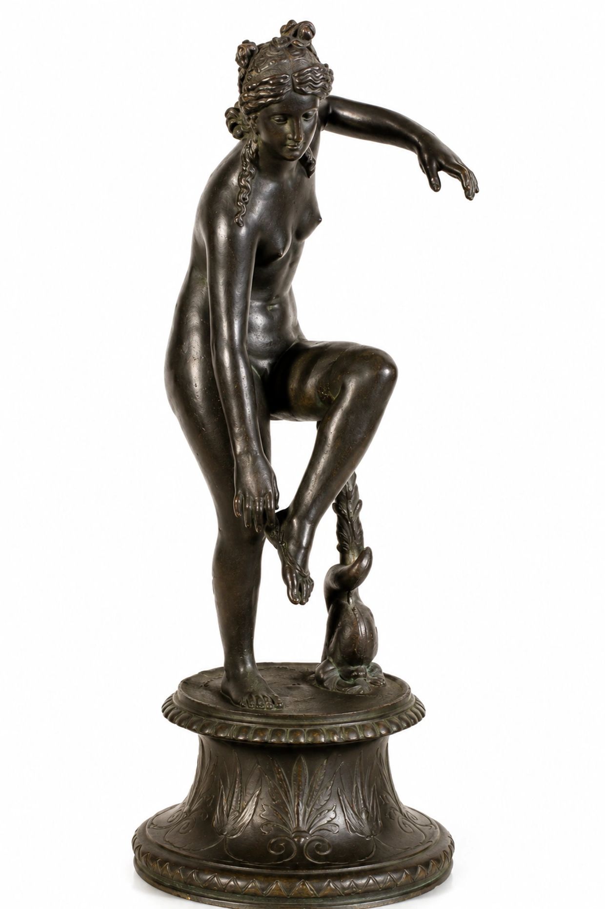 Bronze statue of a woman adjusting her sandal, standing on a decorated base.