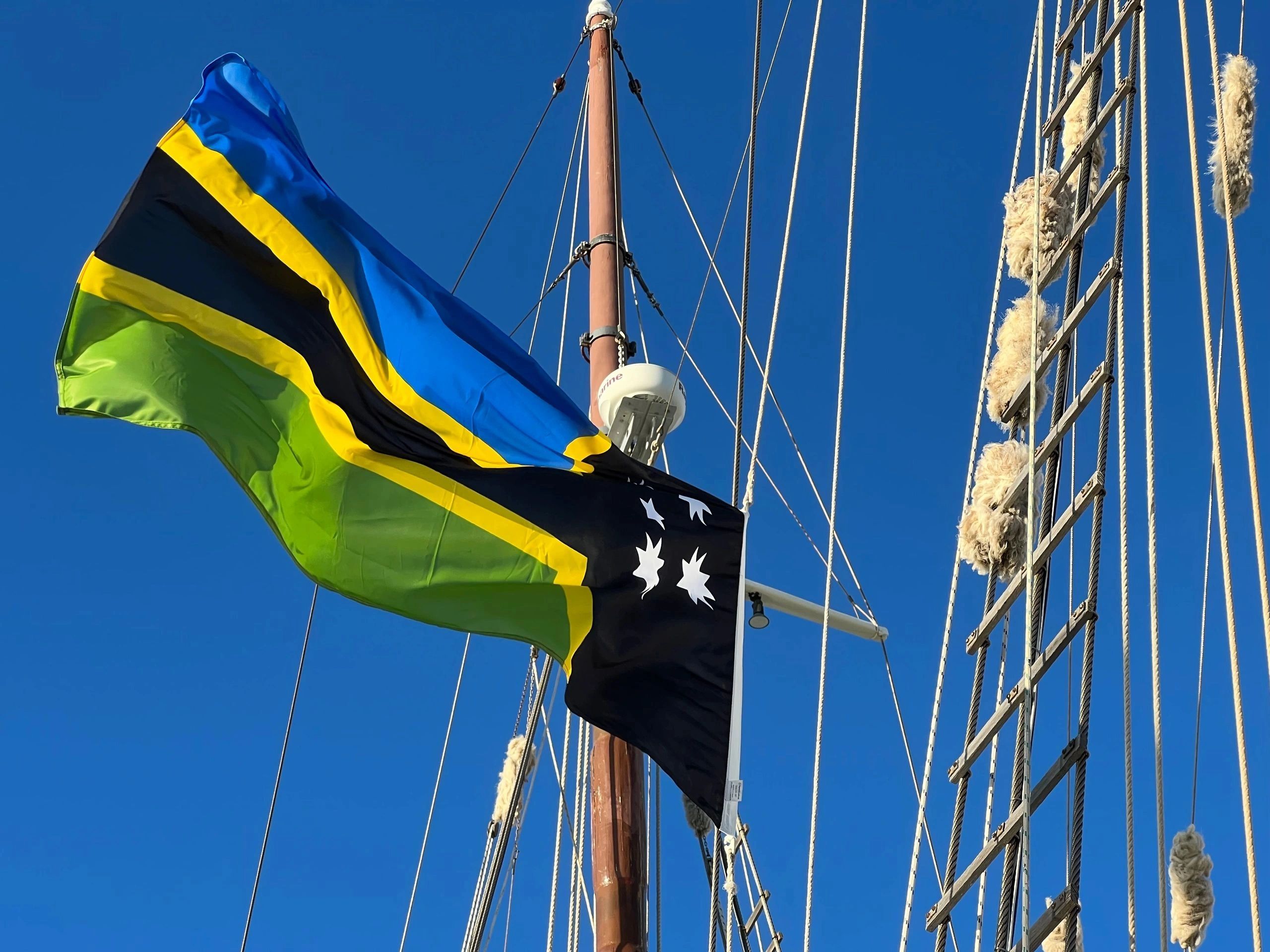 Sailboat mast with a colorful flag against a clear blue sky.