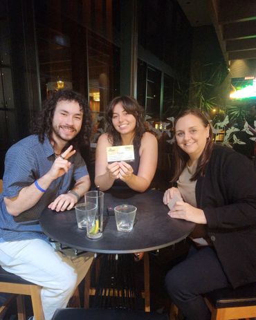 Three friends sitting at a table, smiling and holding a card.