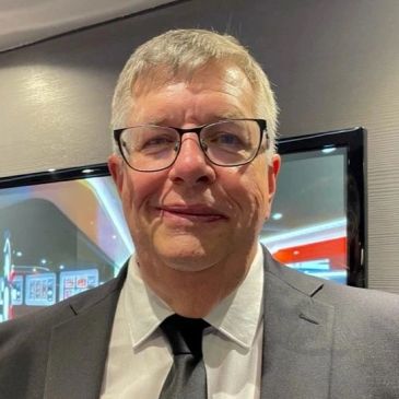 A man in a dark suit and glasses smiles softly indoors.