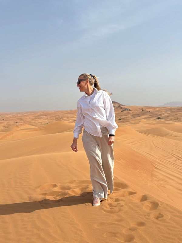 A woman stands in the desert wearing light clothing and sunglasses.