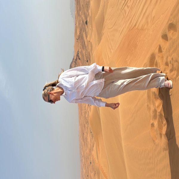 A woman stands in the desert wearing light clothing and sunglasses.