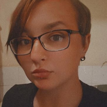 Person with short hair, glasses, and piercings wearing a black shirt.