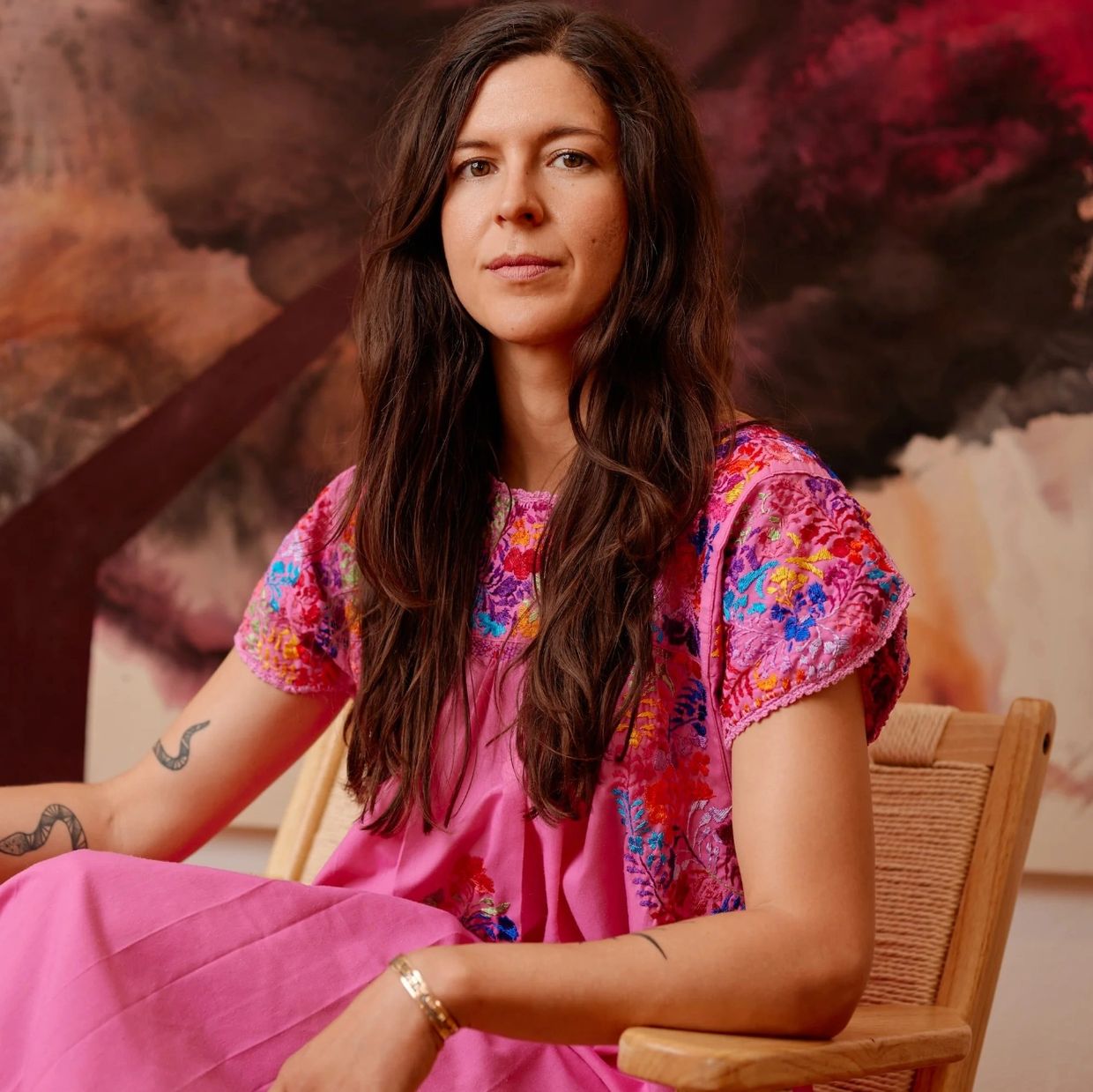 Woman in a vibrant pink embroidered dress sits on a wooden chair with a serene expression.